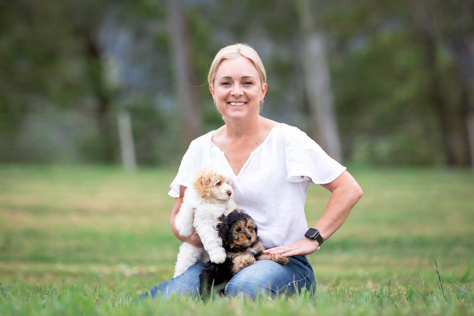 Sarah, owner of Waterhill Cavoodles kneeling wearing a white shirt and jeans holding a white cavoodle puppy and a black cavoodle puppy with a caramel face. Sarah is located in a field on her Sunshine Coast hinterland property.