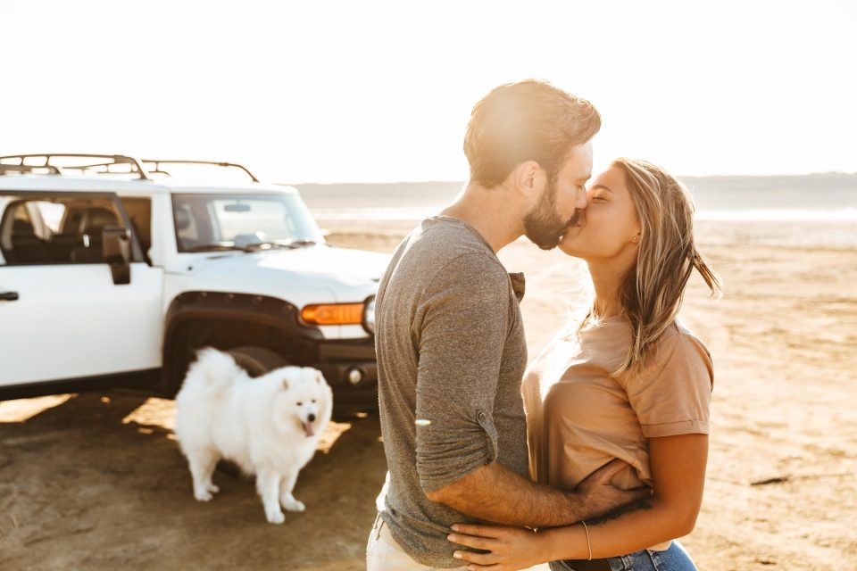 Young couple kissing on beach honeymoon with pet dog sitting by white jeep